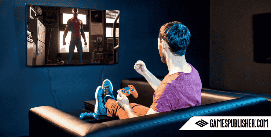 An adult male gamer sitting on a black leather couch, excitedly playing Marvel’s Spider-Man on a PS4, shown on a large wall-mounted TV screen. The scene features Spider-Man standing in an apartment setting. GamesPublisher.com logo appears in the bottom corner.